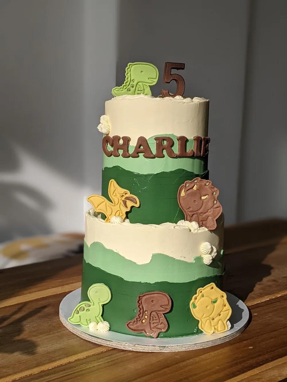 two-tier dinosaur cake in layered greens with dinosaur cookies and a chocolate name