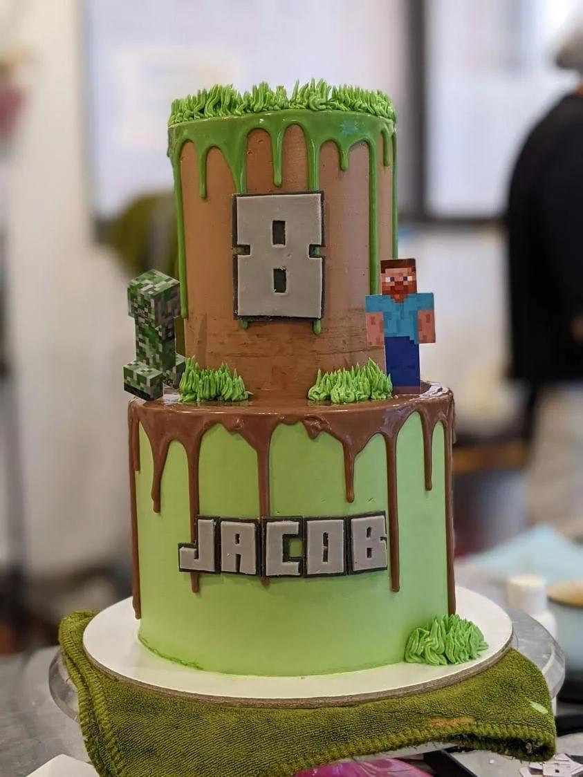 two-tier minecraft cake with chocolate and green drips, piped grass and a block-letter name