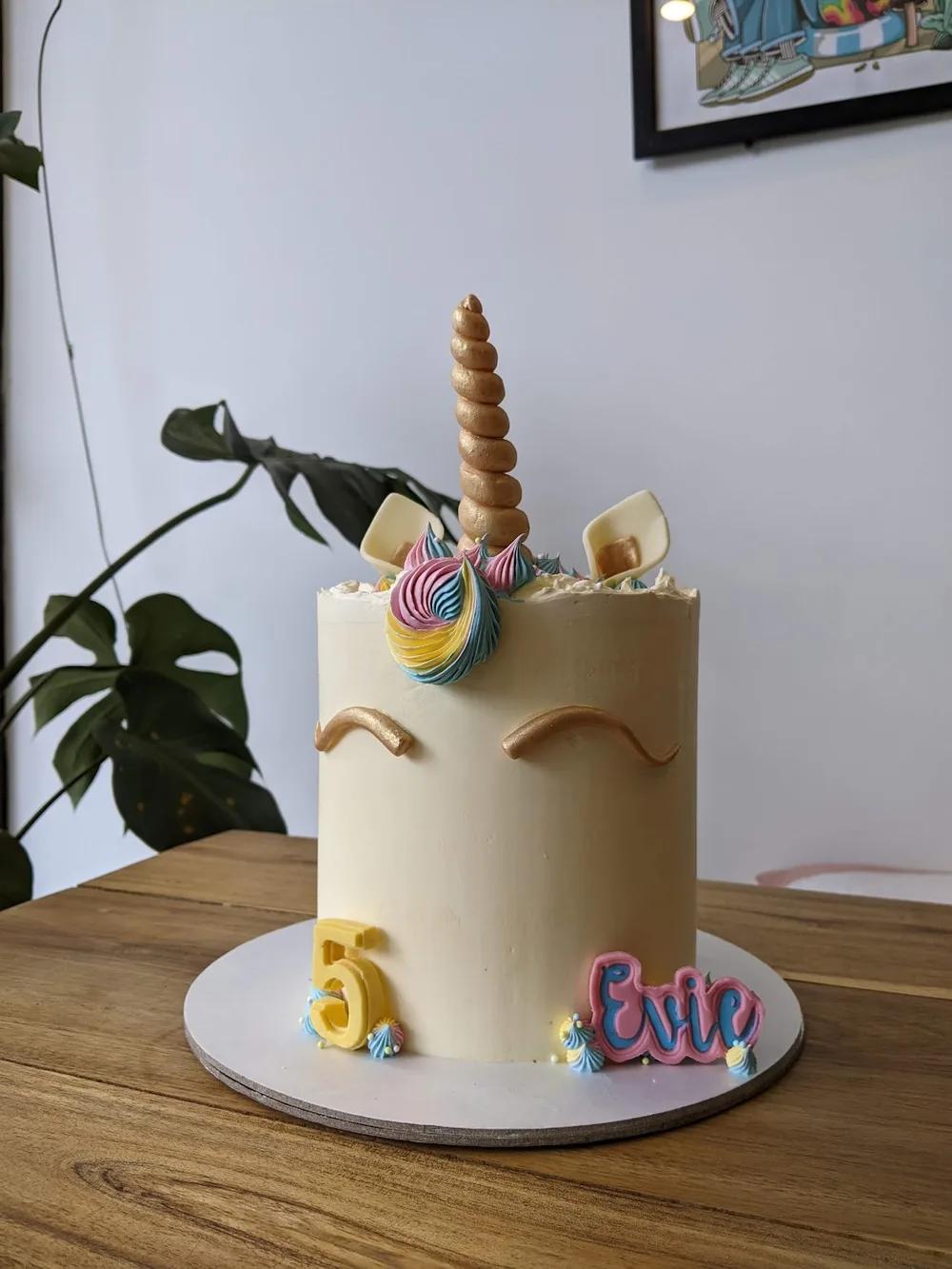 cream unicorn cake with a gold horn, closed gold eyes and a rainbow rosette